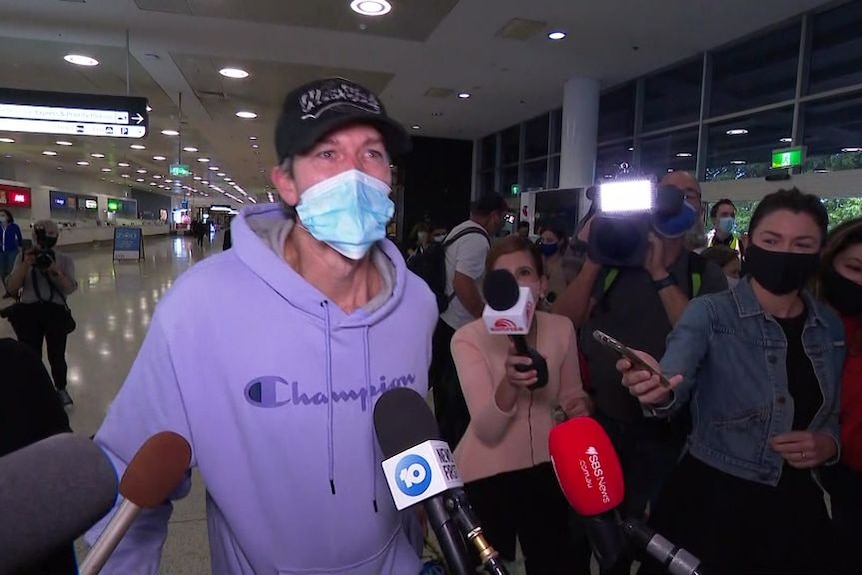 A man wearing a purple jumper and a face mask is interviewed by news crews at Sydney Airport.