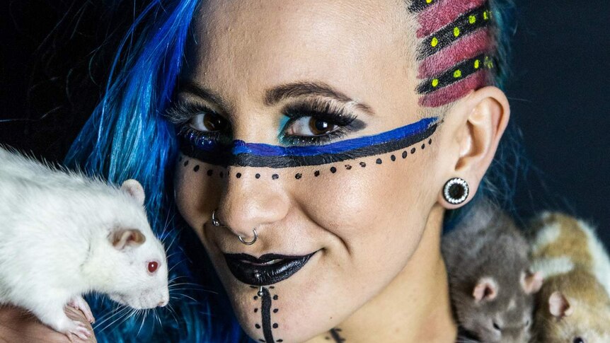 Wrestler Erika Reid, with blue face-paint, holds a rat while two other rats rest on her left shoulder.