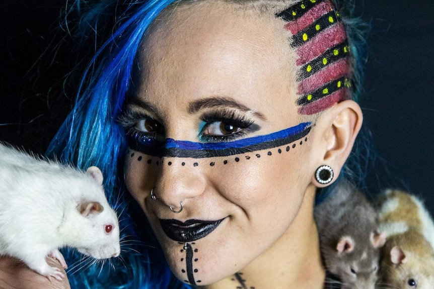 Wrestler Erika Reid, with blue face-paint, holds a rat while two other rats rest on her left shoulder.