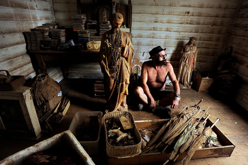 A man sits without a shirt surrounded by his flood and mud damaged possessions