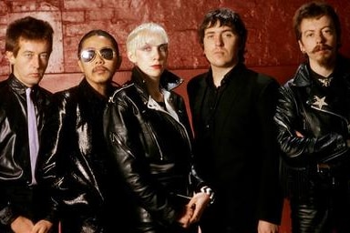 A band poses in front of a brick wall in dark jackets.