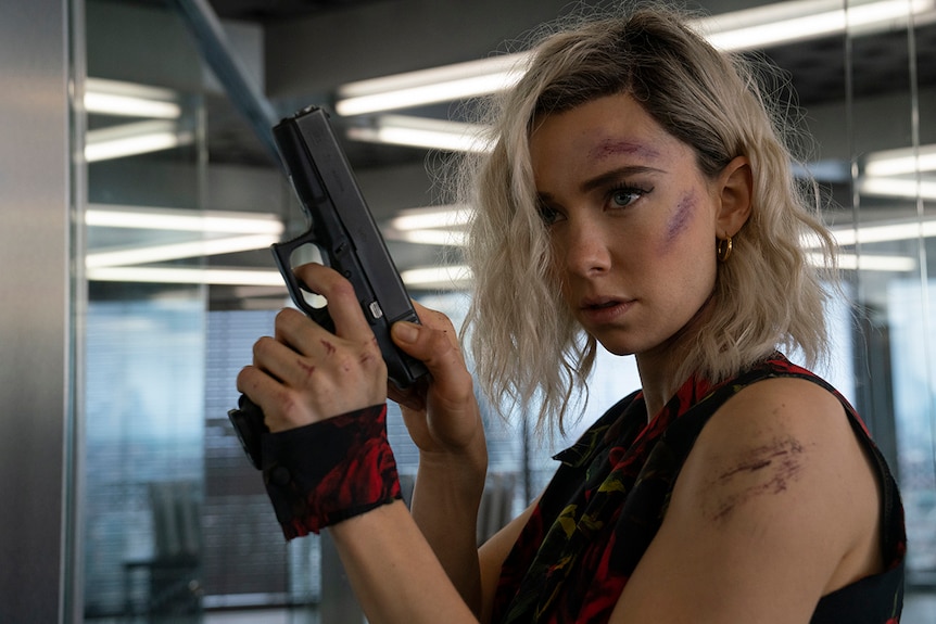 A woman with various combat scratches and concentrated expression cocks handgun in empty glass filled office space.