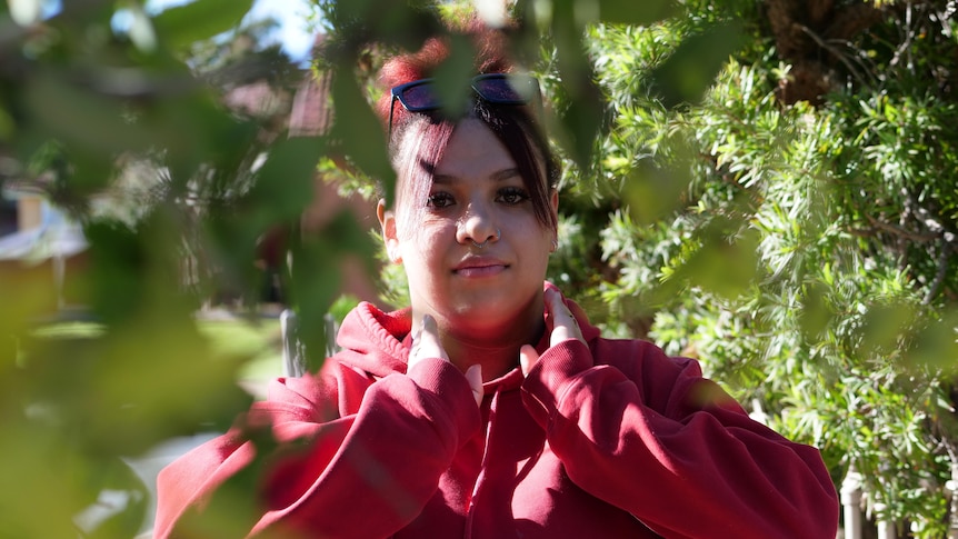 A young woman wearing a red hoodie stands outside, surrounded by tree leaves.