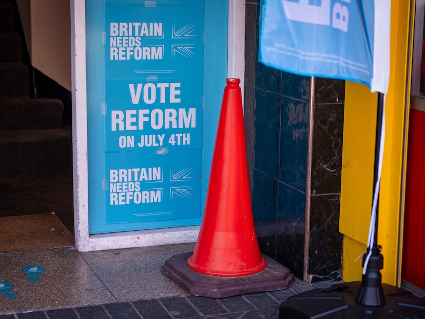 A blue and white card saying 'Vote Reform on July 4th' is pasted on a window with a traffic cone in front of it