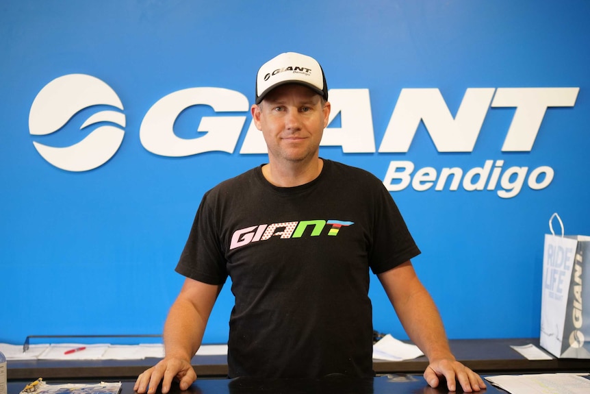 A man wearing a baseball cap and T-shirt stands behind a counter in front of a large blue sign that says Giant Bendigo.