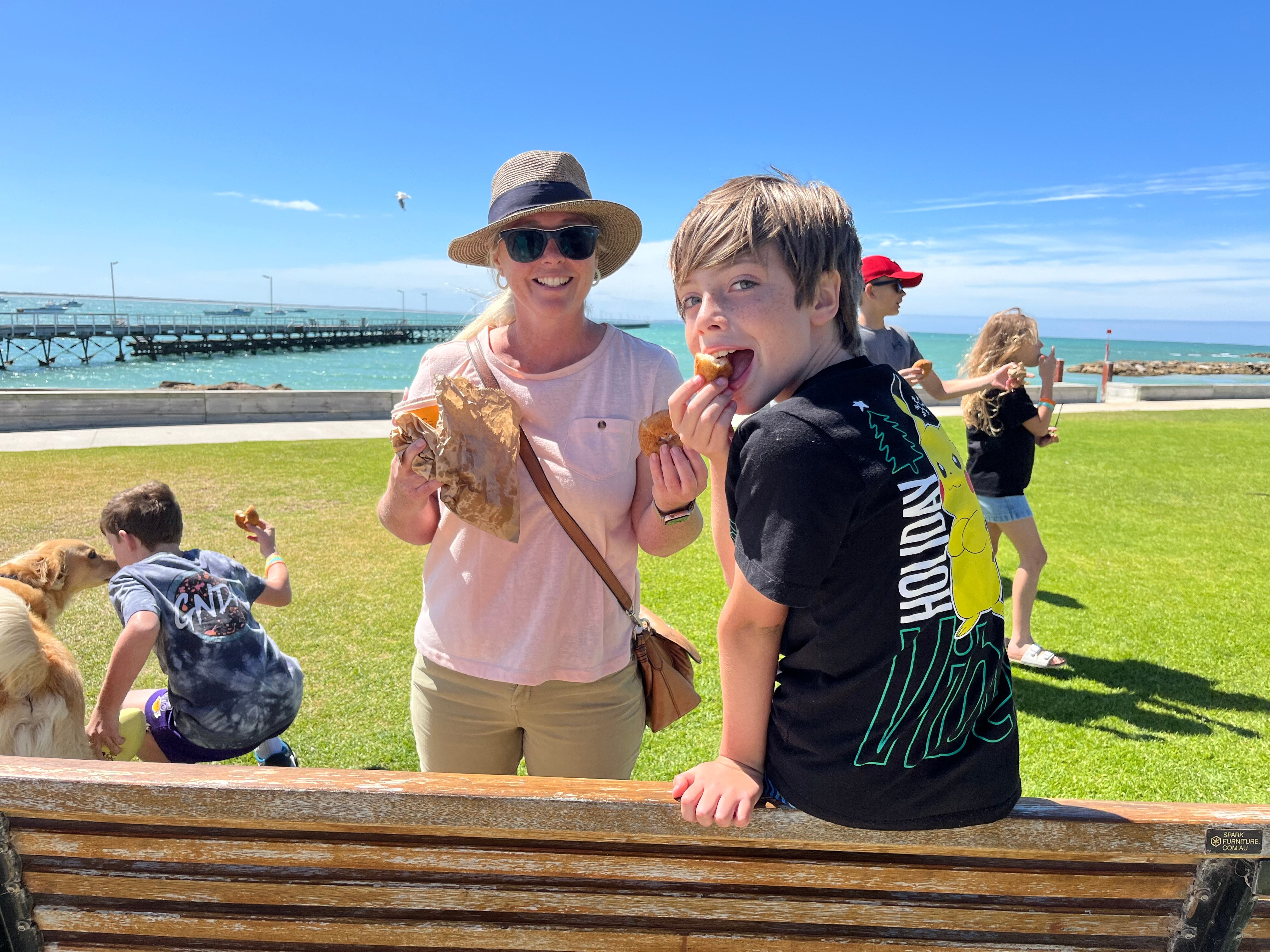 A woman standing and a boy sitting on the back of a bench holding doughnuts with the sea and a jetty behind