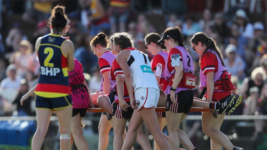 A group of AFLW players surround a teammate on a stretcher who is being taken off the ground.