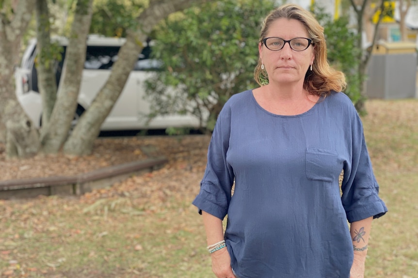 A woman in a blue shirt stands in a backyard. She looks forlorn.