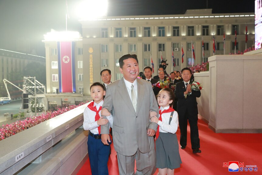Kim Jong Un walks along a red carpet with a child clinging to each arm.