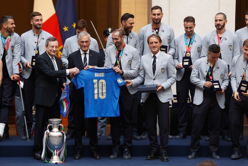 The Italian Prime Minister holds the edge of a signed Italy soccer jersey with his name 'Draghi' on it, as team members watch.