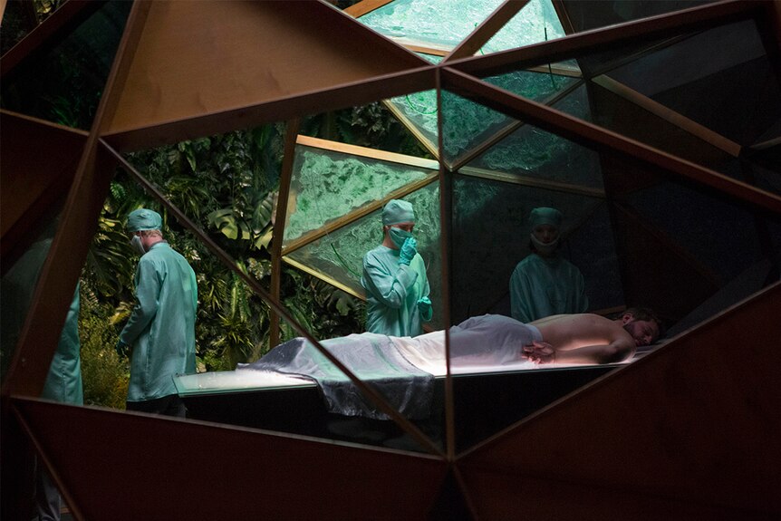 Still image of Logan Marshall-Green lying on a surgeon's table in a geometric room with glass, plants and people in scrubs.