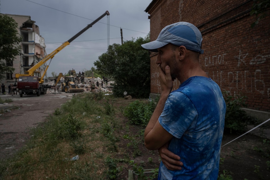 A frightened man in a blue t-shirt waits with his left hand over his mouth as rescuers search the nearby rubble of a building.