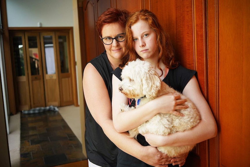 Sally Hardy hugs her teenage daughter Adelaide Hardy and their dog standing in a hallway in their house in Brisbane.