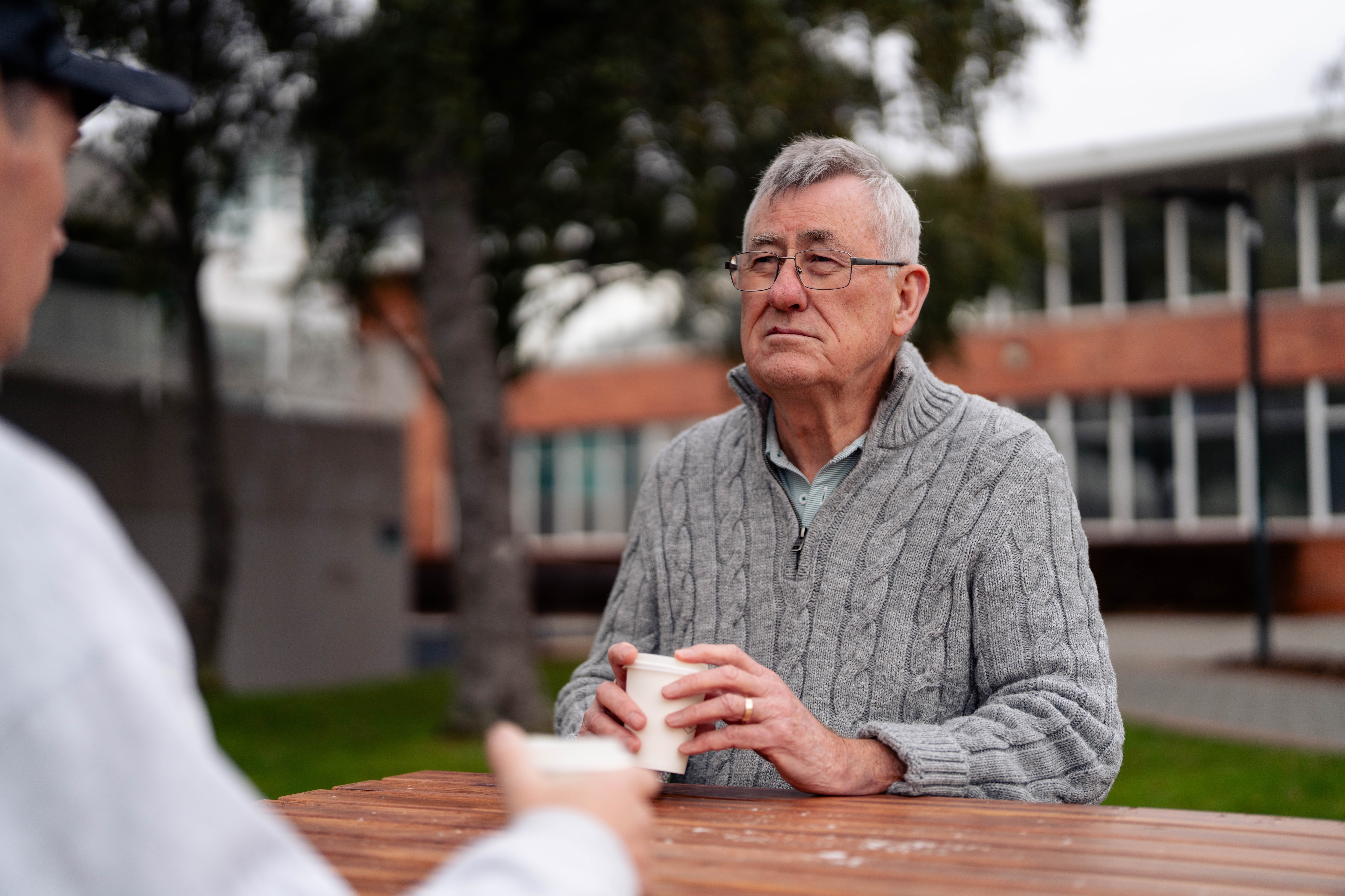 Image taken over Shannon's shoulder, looking to Brendan, wearing a grey quarter zip, sitting at a table with a coffee cup..