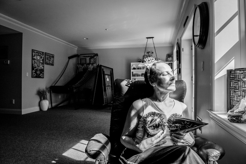 Laurel Borowick sits in an armchair, with party hats placed on breast, letting the sun shine on her face, with her eyes closed.