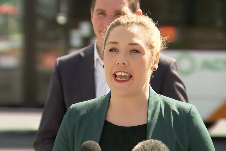 A woman in a green jacket speaks as a bus drives past.