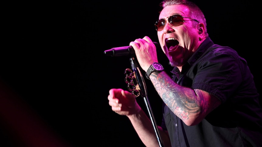 Steve Harwell wearing sunglasses singing into a microphone, his arm is tattooed and he is wearing a black shirt and watch