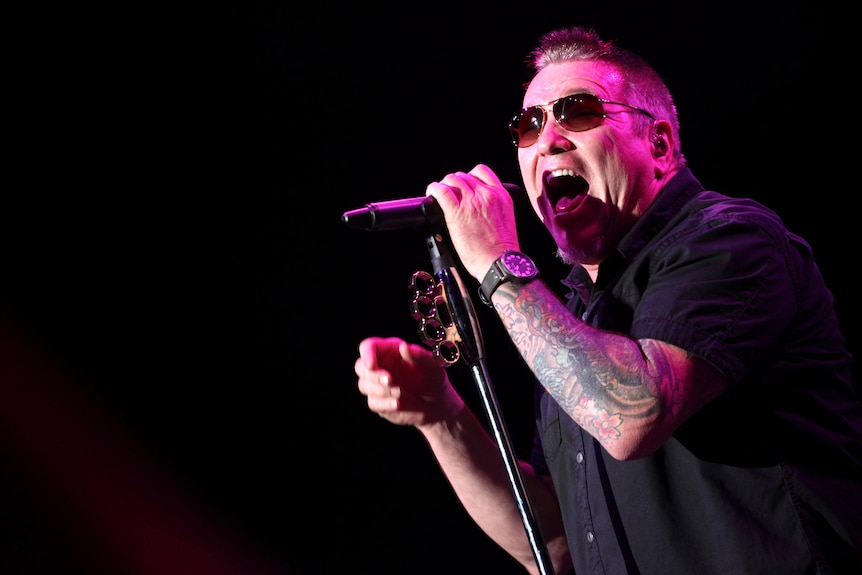 Steve Harwell wearing sunglasses singing into a microphone, his arm is tattooed and he is wearing a black shirt and watch