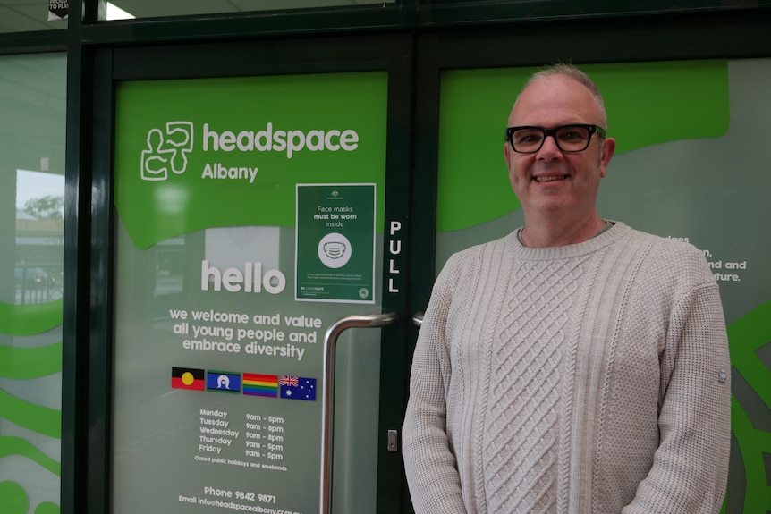 Man in a white knit cardigan stands smiling in front of a green door bearing a pride flag.