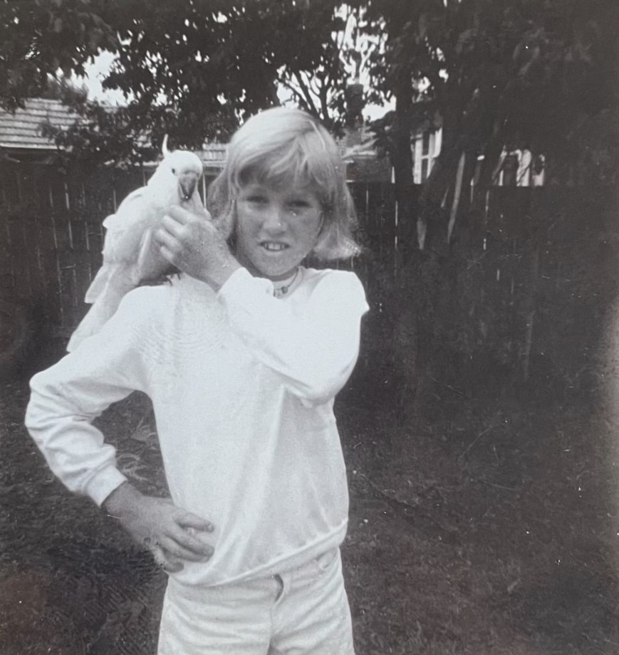 Black and white photo of a blonde boy dressed in white with a cockatoo on his shoulder.