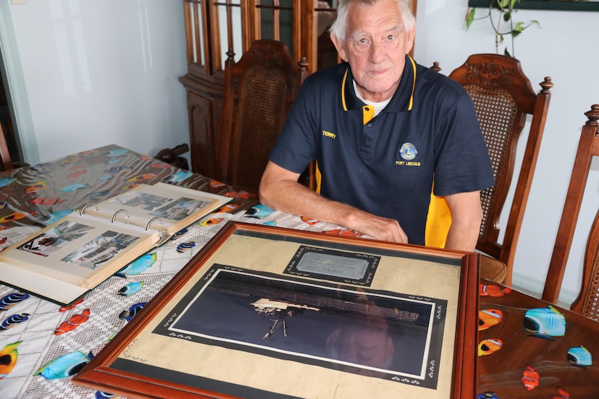 Old man sitting at table looking at large frame print of boat and name tag
