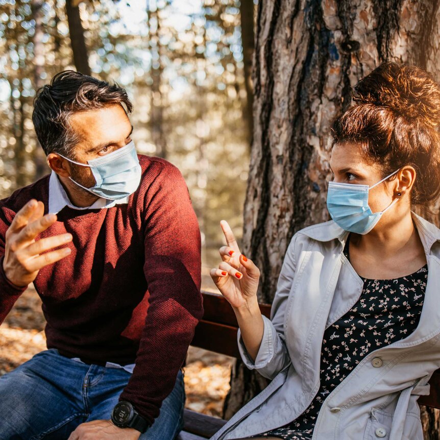 A man and a woman sitting on a bench in a park both wearing face masks