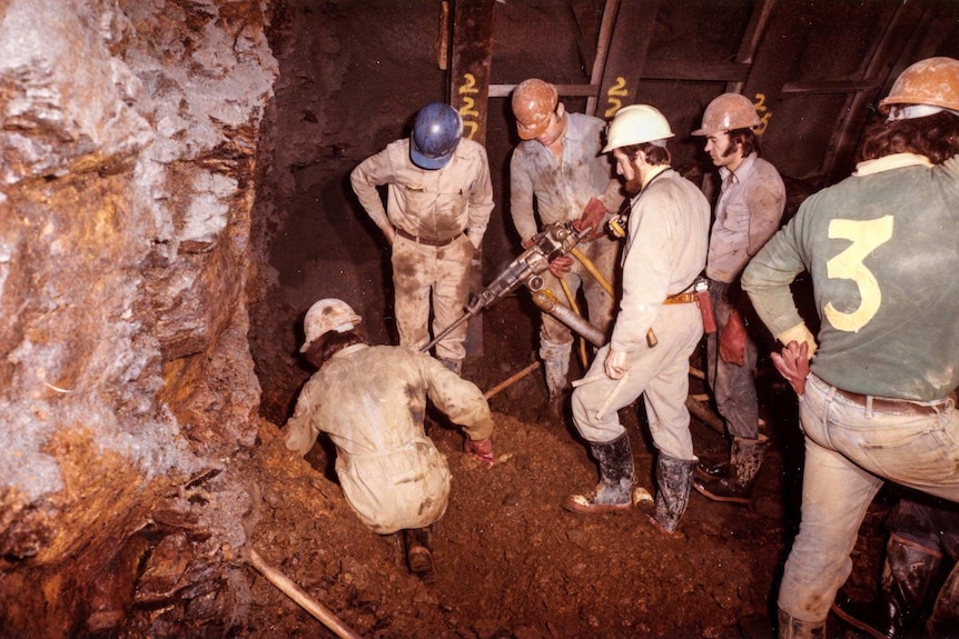 Six men, some with shoulder length hair, sideburns or beards, work underground with a drill