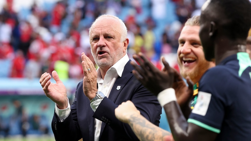 A Socceroos coach claps as he stands near the pitch and players to his right laugh in celebration after a game.