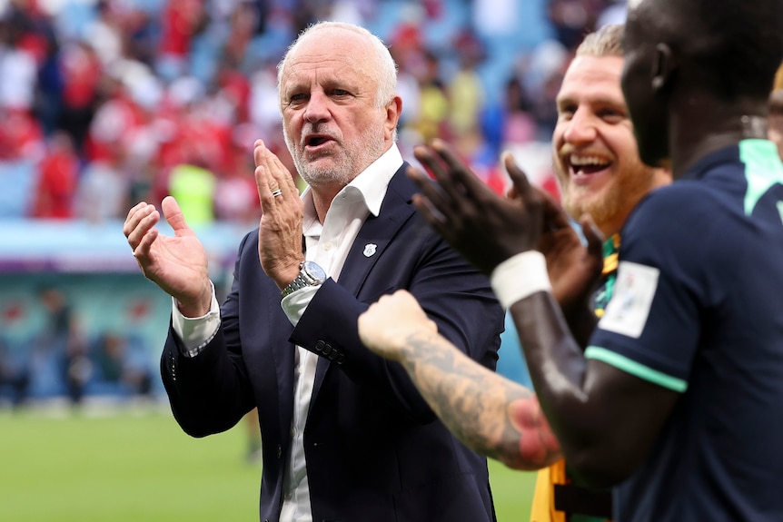 A Socceroos coach claps as he stands near the pitch and players to his right laugh in celebration after a game.