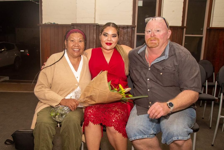 Three people sit smiling with their arms around each other.