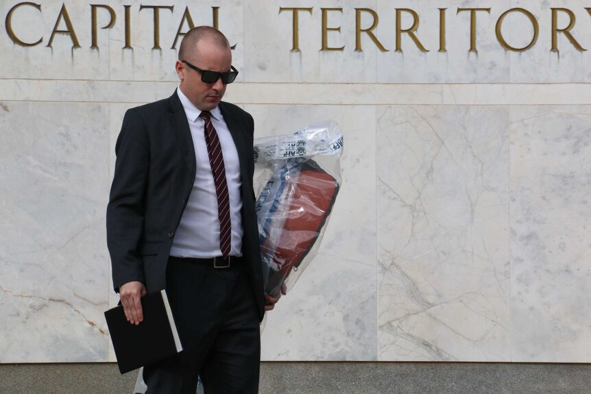 A police officer in a suit carries an AFP evidence bag containing a rifle case.