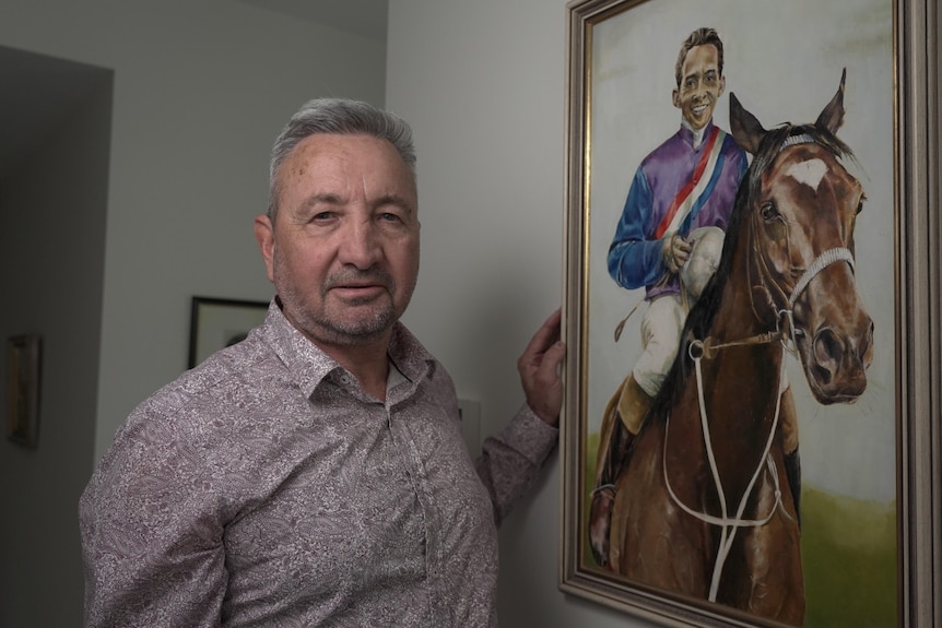 A middle-aged man stands in front of a painted portrait of a jockey on a horse