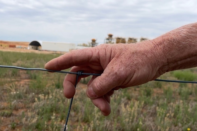 A close up of an older fair-skinned man’s hand, Kevin, rests on a wire fence with a mine in the background.
