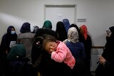 a child rests in her mother's arm as a group of women line up outside a doctor's room