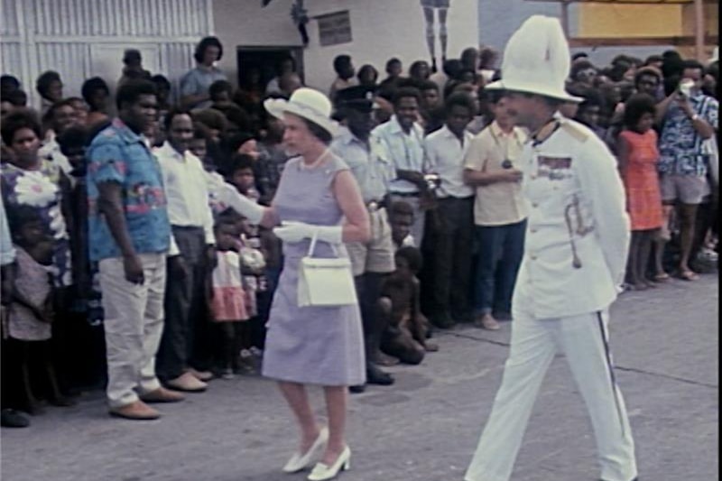 Queen in purple dress and white hat walks past crowd, with man in white soldier uniform.