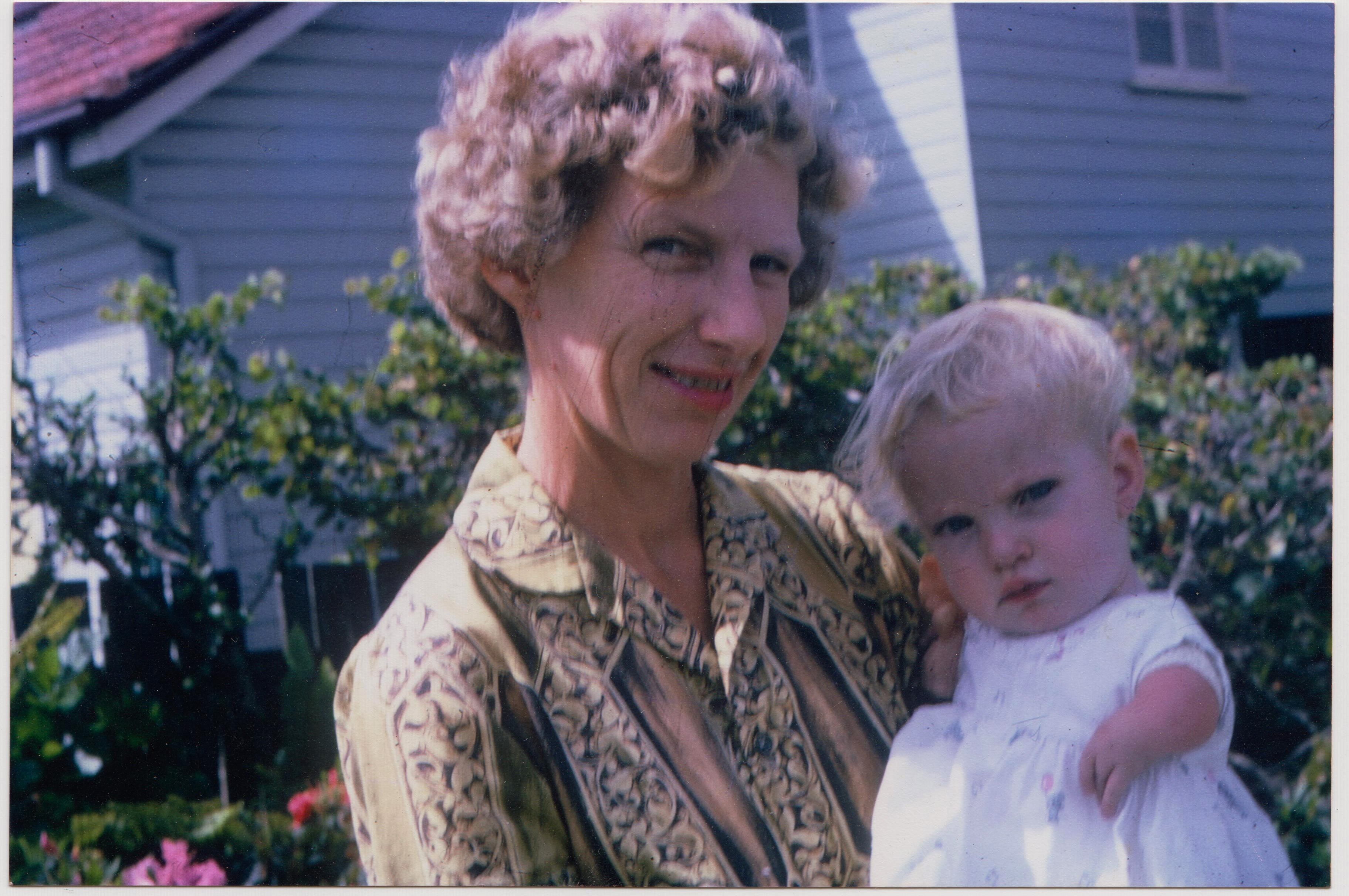 A woman smiles in front of an old Queenslander holding a young baby in a frilly dress.