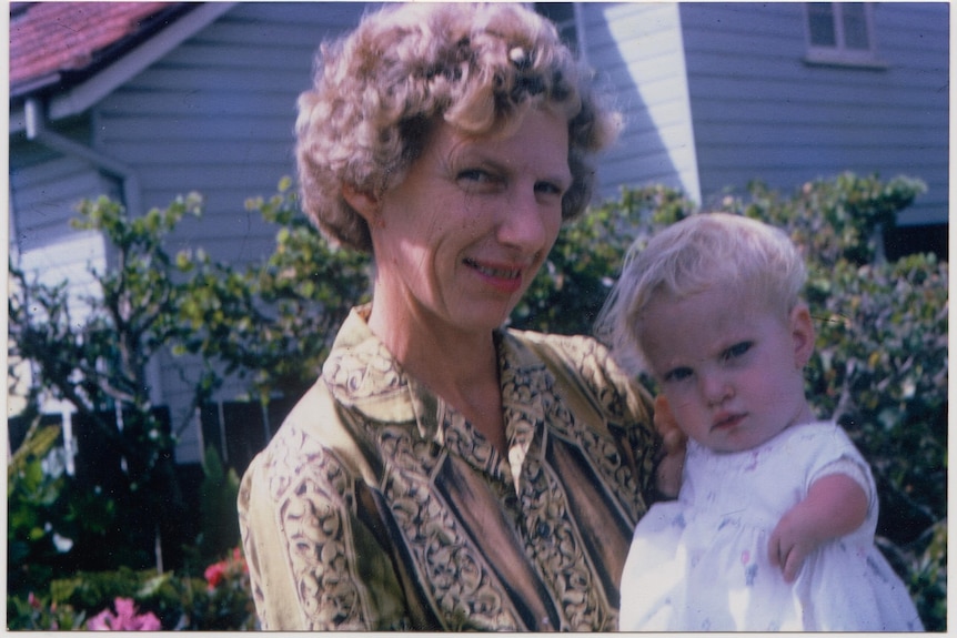 A woman smiles in front of an old Queenslander holding a young baby in a frilly dress.