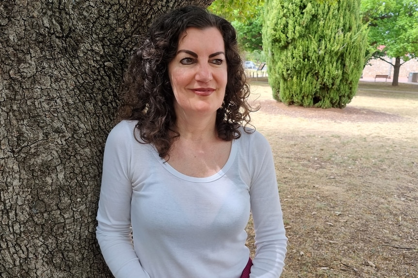 A woman with brown curly hear wearing a white top stands in front of a tree looking into the distance.