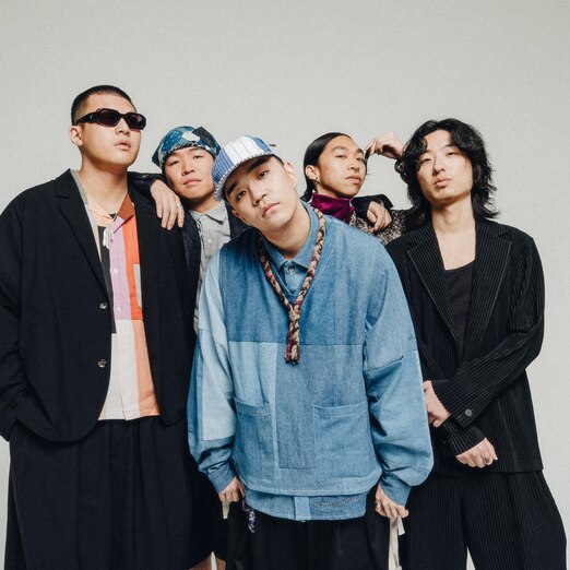 Five Korean men stand in front of a grey background in streetwear and one is wearing sunglasses