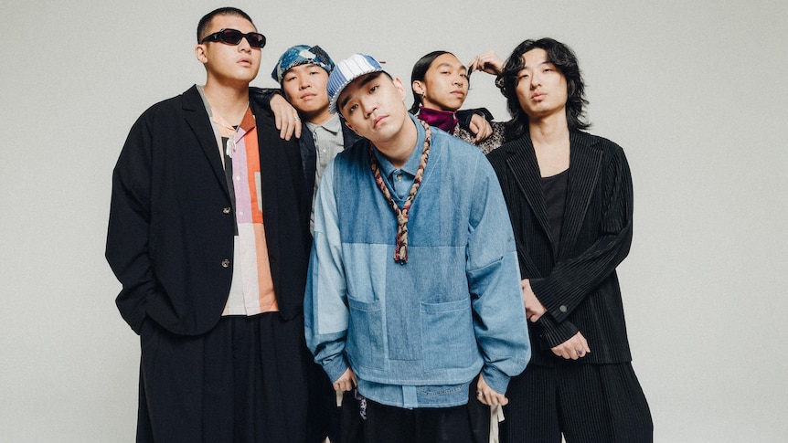 Five Korean men stand in front of a grey background in streetwear and one is wearing sunglasses