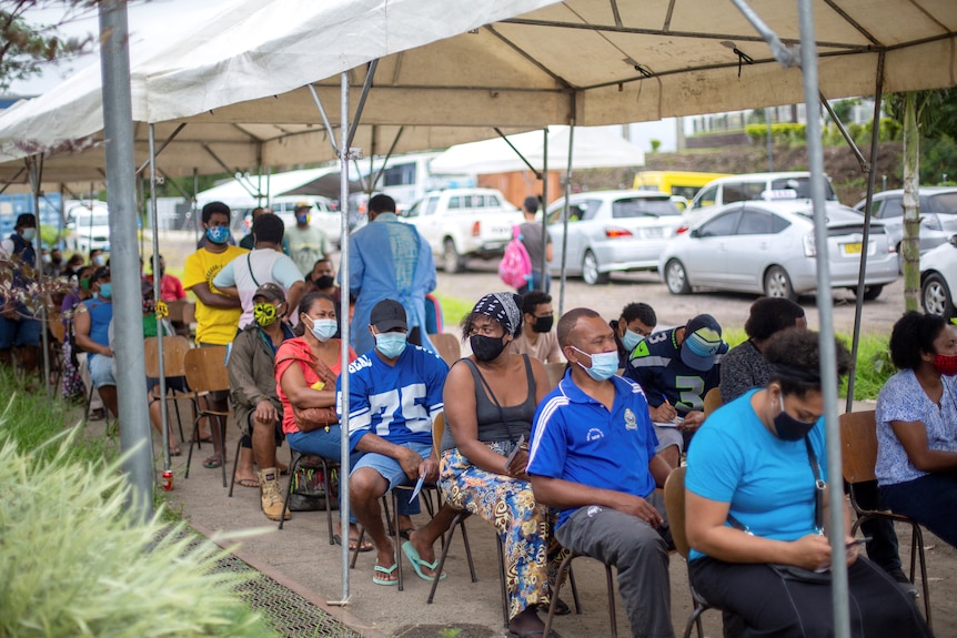 Fijians sit in a line of chairs under a tarpaulin as they wait to receive a dose of the AstraZeneca vaccine