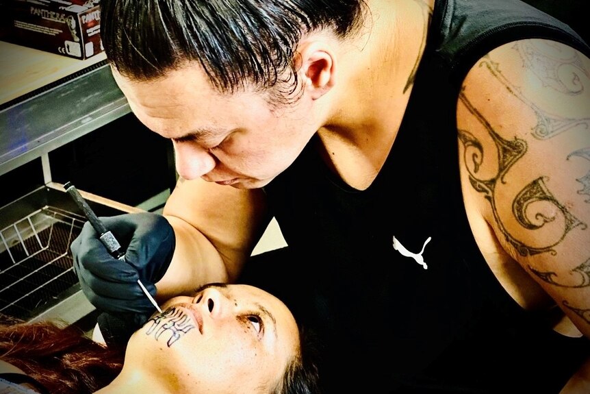 A woman lies on her back as a man bends over to tattoo her chin.