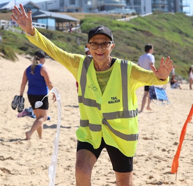 Nancy usando um colete de alta visibilidade para correr no parque sorrindo com as mãos para cima correndo na praia. 