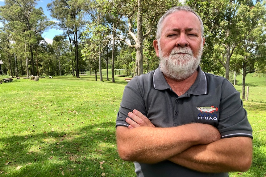 A bearded man looks seriously at the camera, standing with his arms crossed.