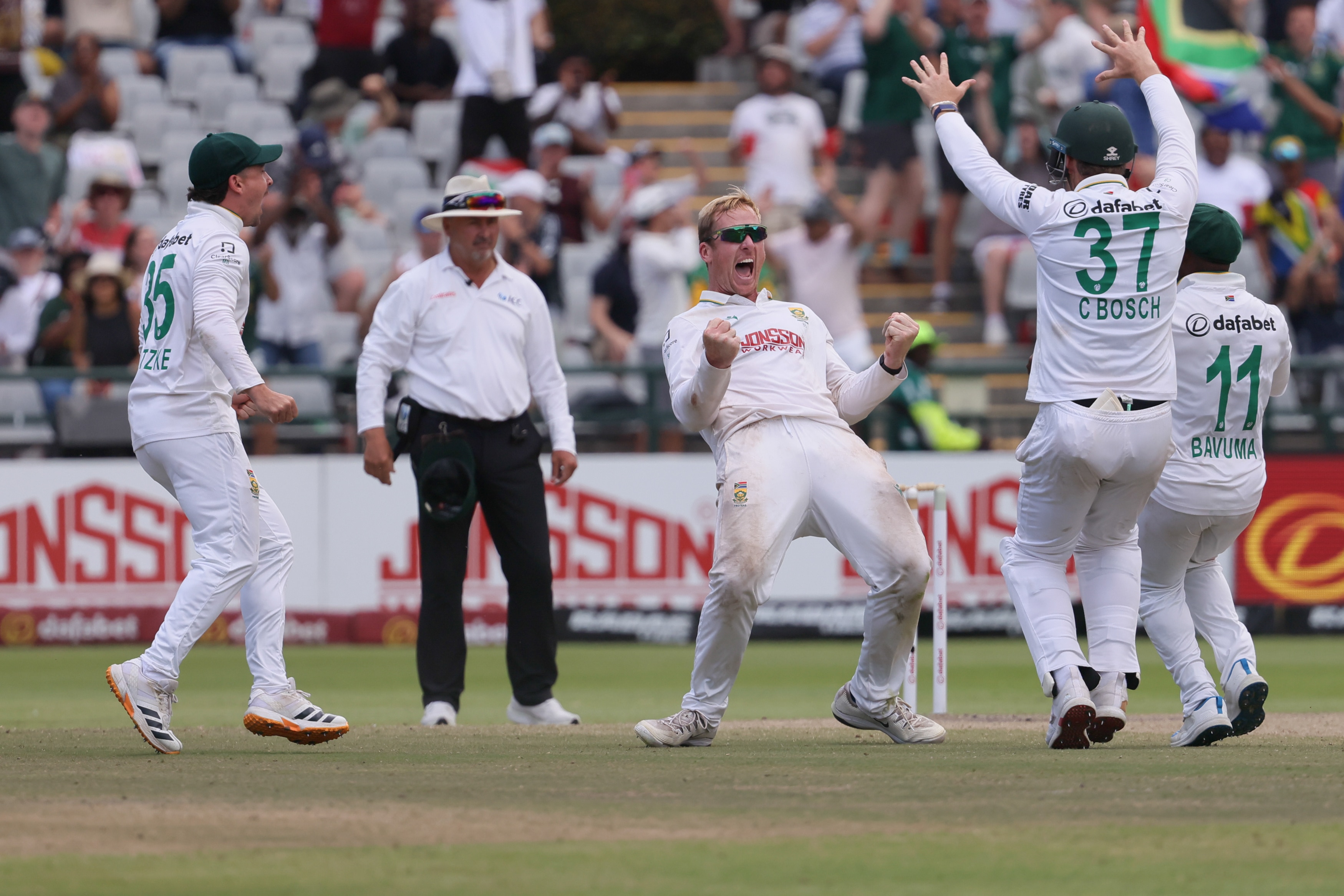 A South African spin bowler yells and pumps both his fists in celebration as his teammates run in after a wicket.