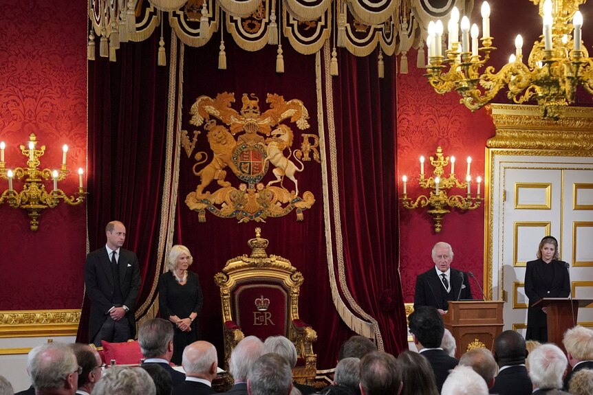 Prince William and Camilla on one side of an ornate throne as King Charles III speaks in front of a crowd. 