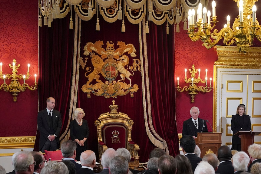 Prince William and Camilla on one side of an ornate throne as King Charles III addresses a crowd. 