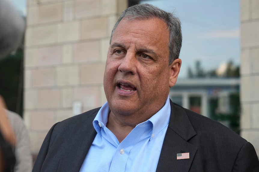 A heavy, middle-aged man with greying hair and wearing a suit speaks in front of a sandstone building.