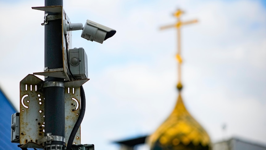 A surveillance camera sits on a utility pole, with the top of a Russian orthodox church in the background.