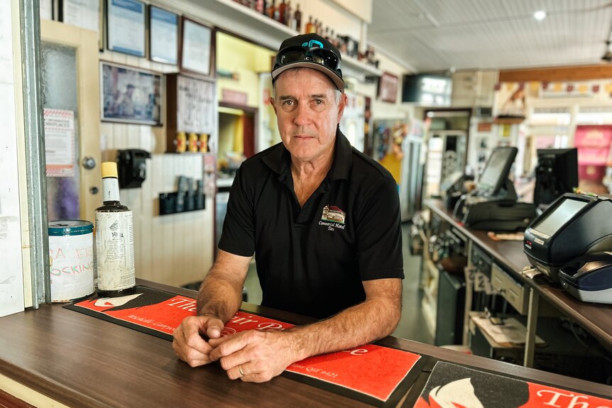 a pub owner leans his elbows on a bar decorated with red bar mats. he's wearing a cap and a black shirt